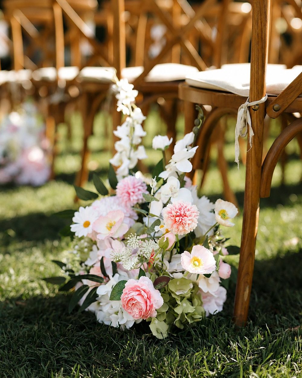 Meadow arrangement of blush and ivory florals set on the grass beside aisle chairs